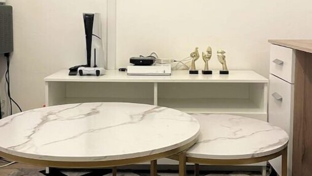 Ein modernes Zimmer mit einem wandmontierten Flachbildfernseher, zwei Marmortische, einem weißen Schreibtisch mit Computer und Dekoration sowie einem gemusterten Teppich.