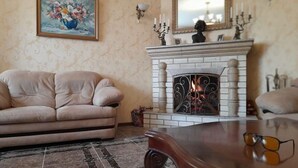 A living room with a beige sofa, a wooden coffee table, a fireplace with a fire, a framed painting on the wall, and a mirror above the fireplace. - Avan Luxury Villa (Arinj)