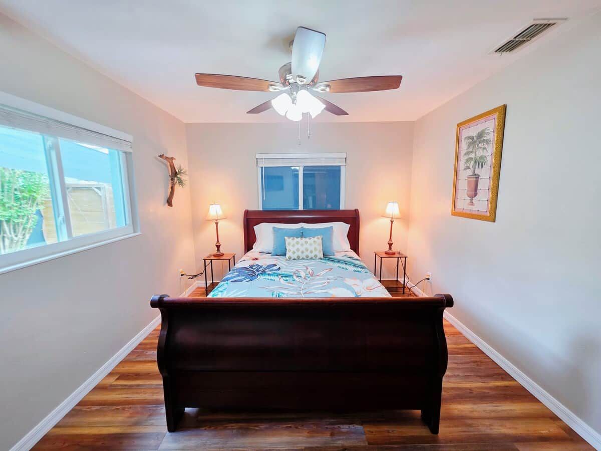 A bedroom with a bed, two bedside lamps, a ceiling fan, a window, and a framed picture on the wall.