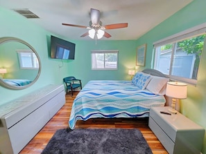 Een slaapkamer met een bed, plafondventilator, nachtkastje, dressoir, stoel en een raam.