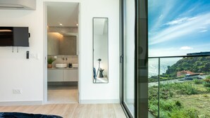 A modern bedroom with a large window, a flat-screen TV, and a bathroom with a mirror and tripod. - Orion Calheta Hills by LovelyStay (Calheta)