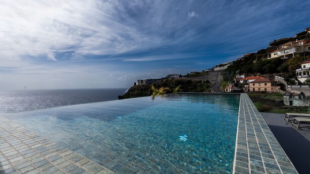 An infinity pool with clear blue water, a tiled deck, and a view of the ocean and hillside buildings. - Orion Calheta Hills by LovelyStay (Calheta)