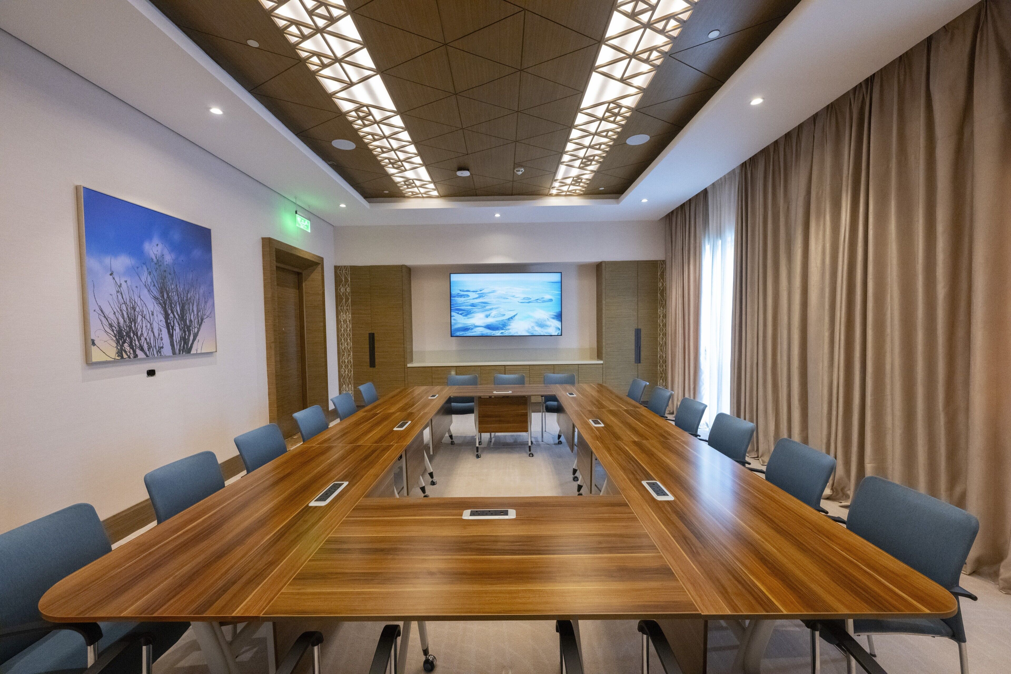 Un salón de conferencias con una larga mesa de madera, sillas azules, una pantalla grande y un cuadro en la pared.