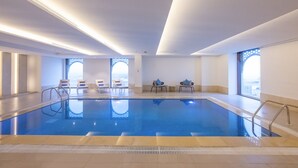 Una piscina interna con acqua cristallina, finestre illuminate dal sole e lettini.