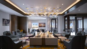 Un'area lounge moderna con divani, poltrone e tavoli rotondi, caratterizzata da un tappeto decorato e un soffitto con lampade a sospensione.