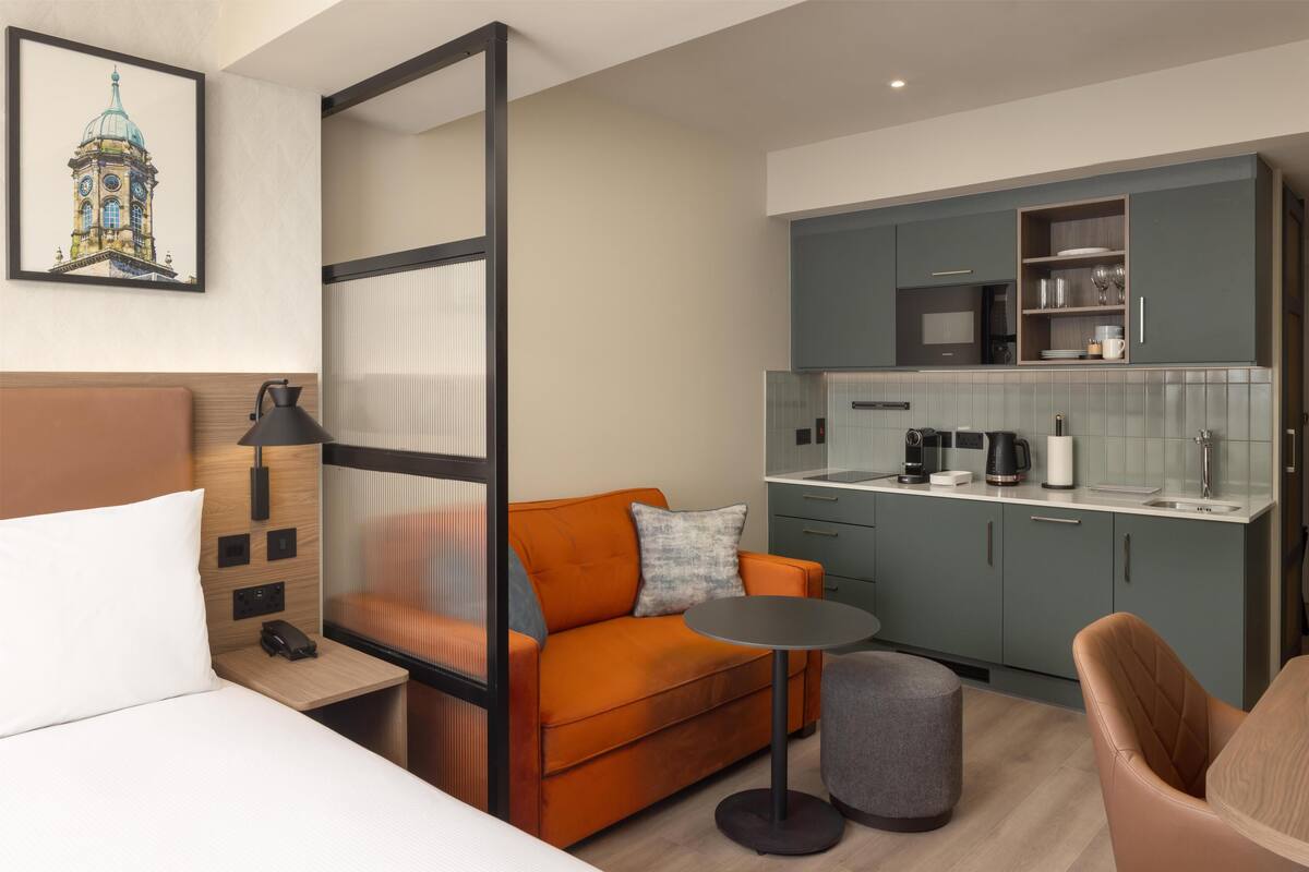 Ein modernes Hotelzimmer mit Bett, orangefarbenem Sofa, kleinem runden Tisch, Küchenzeile und einem Bild an der Wand.