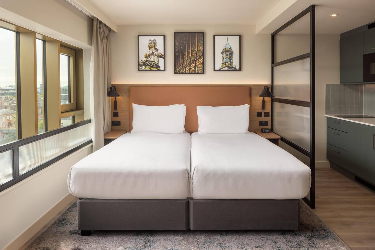 Ein Doppelbett mit Kopfteil, ein Nachttisch, eine Lampe und Blick auf eine Stadtlandschaft durch große Fenster.