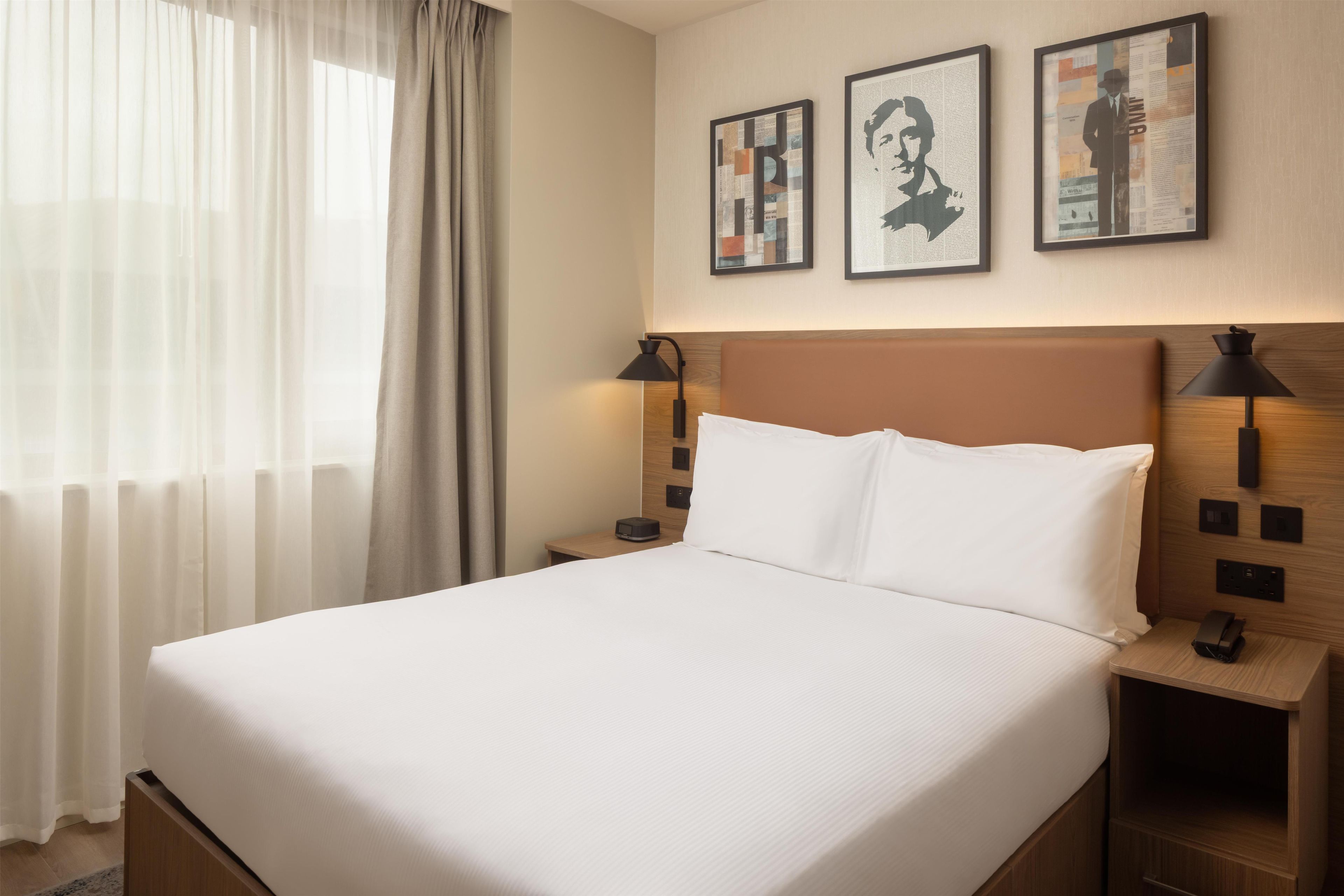 Une chambre d’hôtel avec un grand lit, deux tables de chevet avec des lampes, une fenêtre avec des rideaux légers et trois tableaux encadrés accrochés au mur.