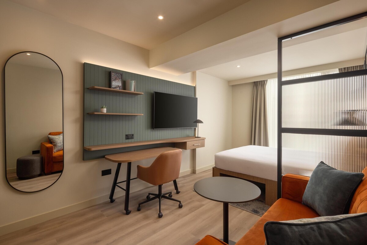 Una habitació d'hotel moderna amb un llit, una taula de treball amb una cadira, una taula petita, un televisor fixat a la paret i una finestra gran.