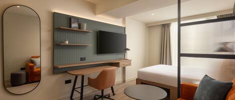 Una habitación de hotel moderna con una cama, un escritorio con una silla, una mesita pequeña, un televisor montado en la pared y un ventanal grande.