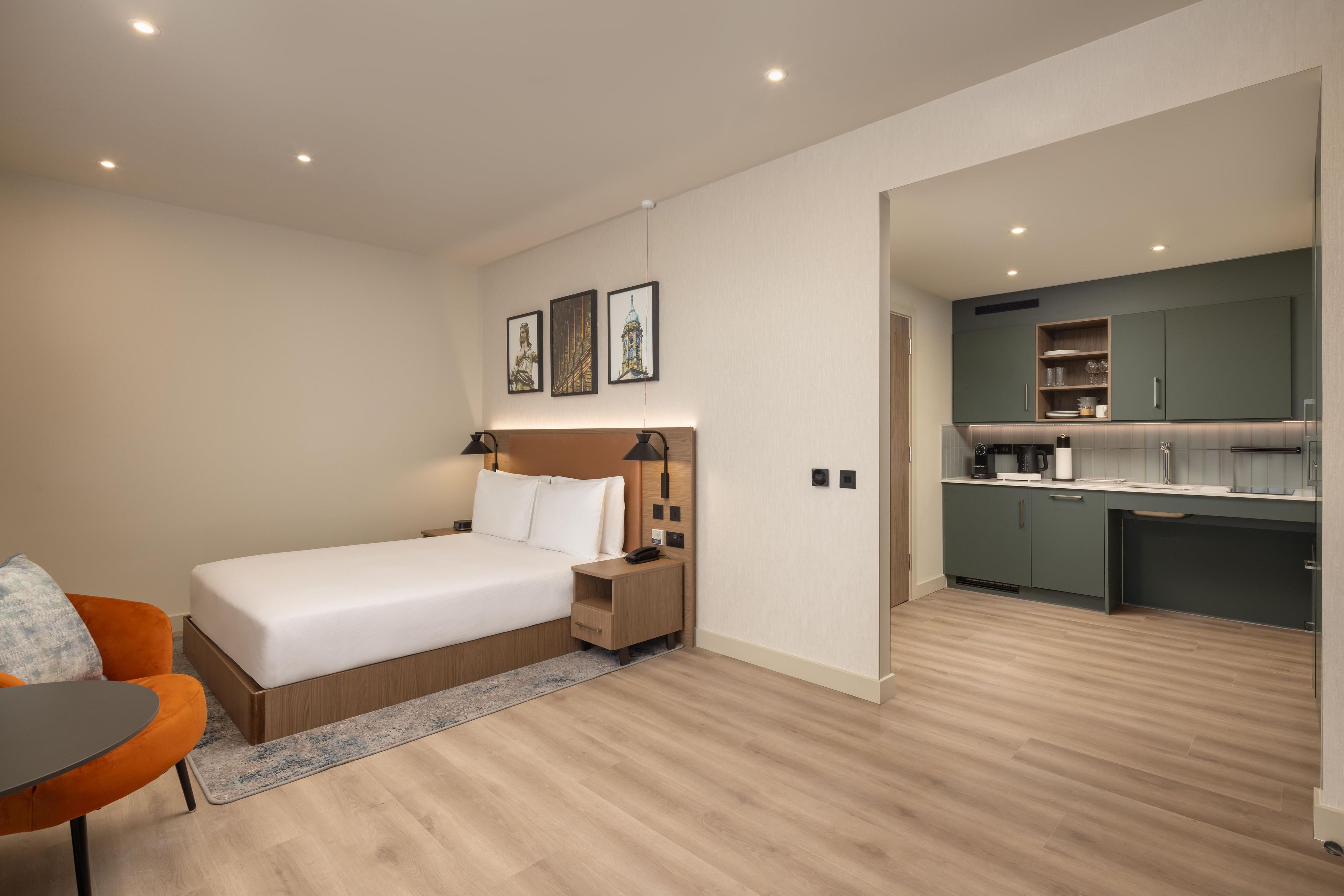 Een moderne slaapkamer met een bed, nachtkastje en een oranje stoel. Grenzend daar aan bevindt zich een kitchenette met groene kastjes en een aanrecht.