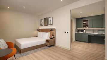 Een moderne slaapkamer met een bed, nachtkastje en een oranje stoel. Grenzend daar aan bevindt zich een kitchenette met groene kastjes en een aanrecht.