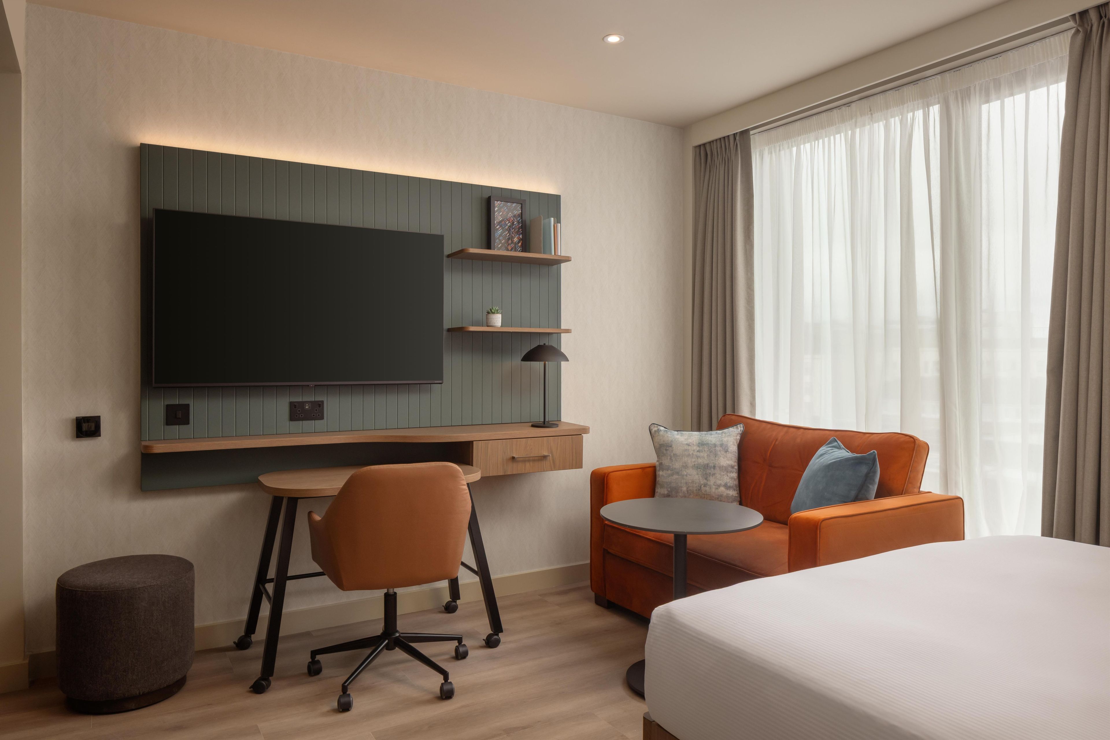 Una habitación de hotel moderna con cama, escritorio, sillón, sofá, televisor y ventana con cortinas.
