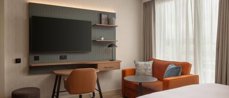Una habitación de hotel moderna con cama, escritorio, sillón, sofá, televisor y ventana con cortinas.