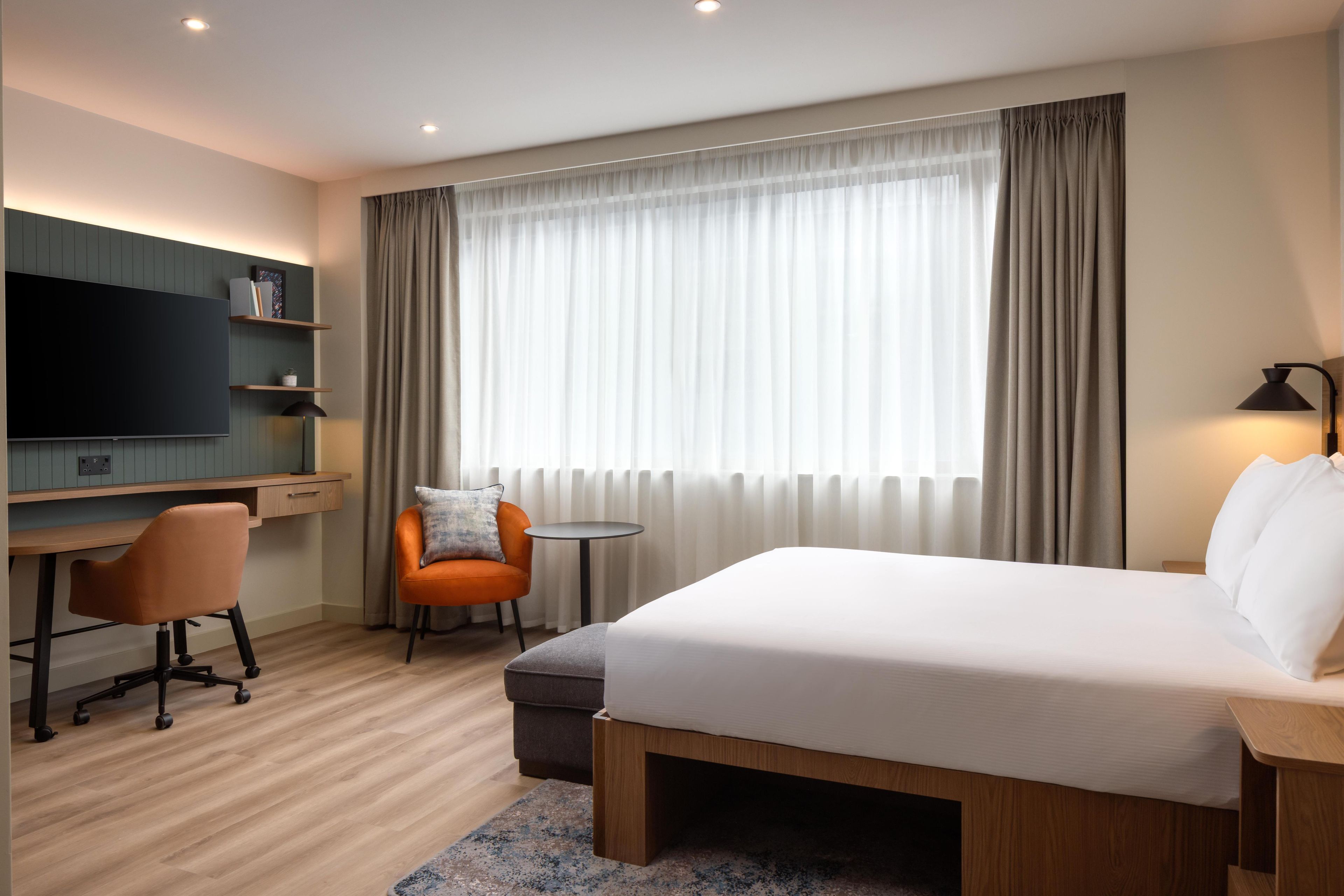 Quarto de hotel com cama, escrivaninha, cadeira, televisão e janela com cortinas.