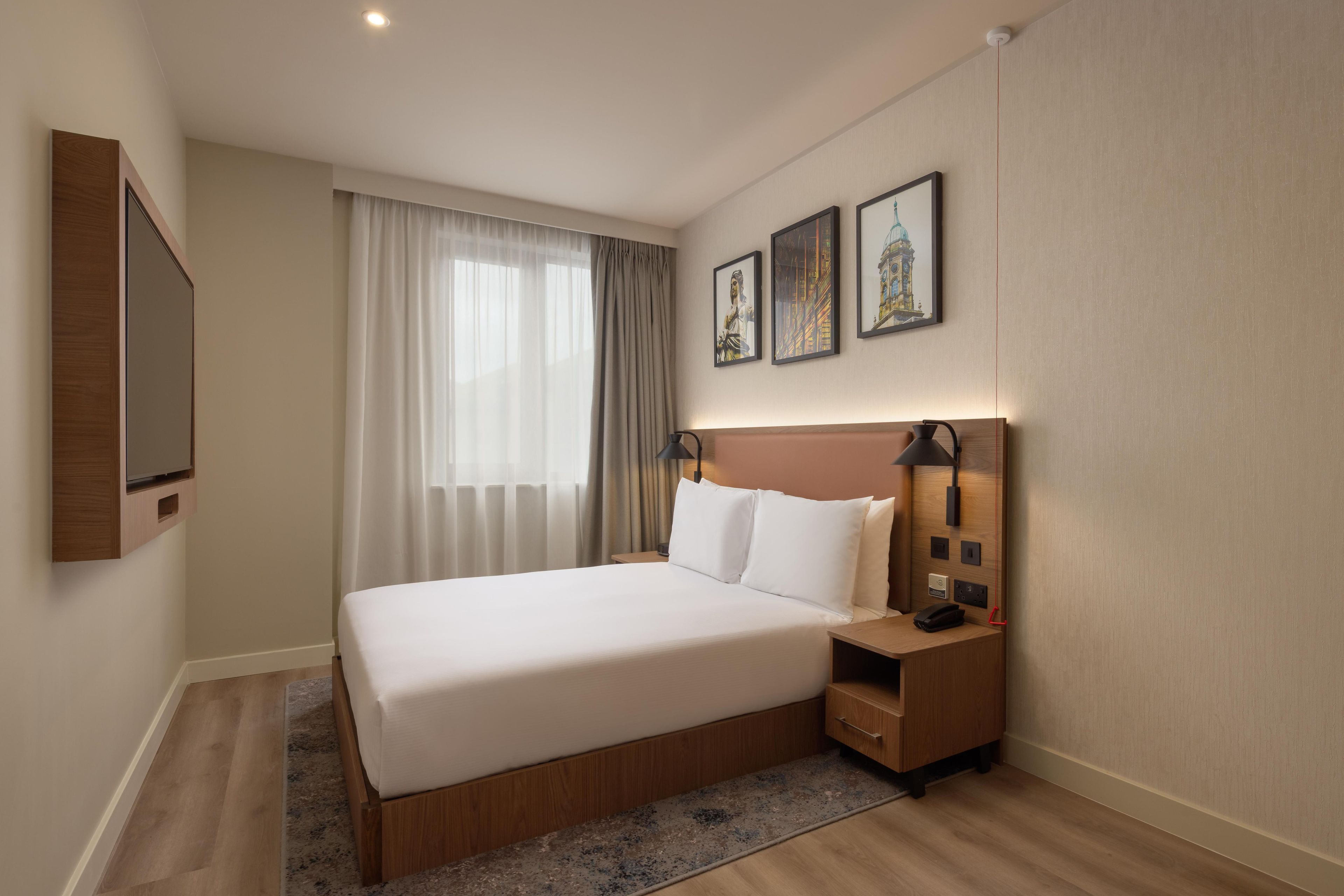 A hotel room with a large bed, a bedside table with a lamp, a flat-screen TV, and three framed pictures on the wall.