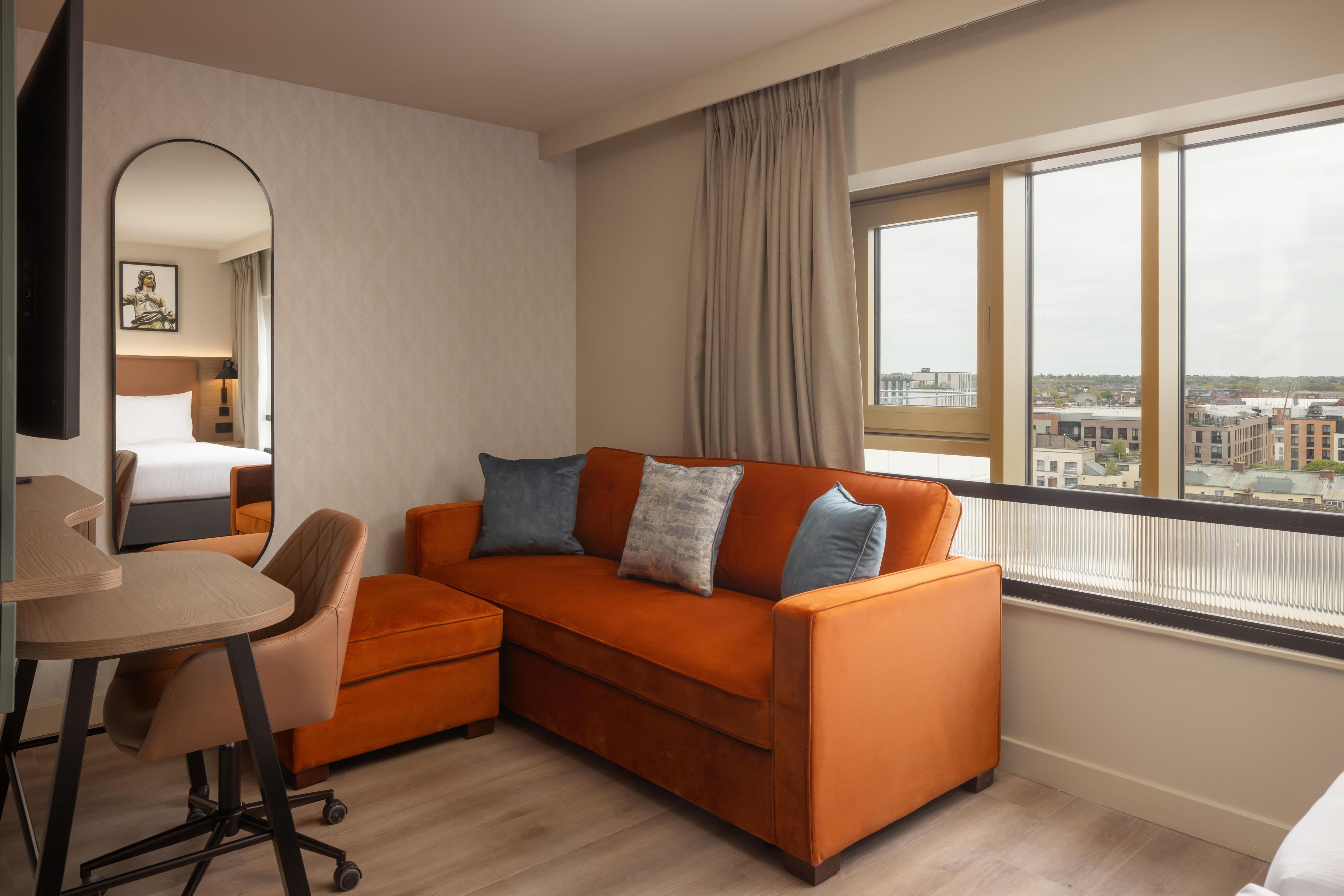 Et moderne hotelværelse med en sofa, et skrivebord og et stort vindue med udsigt over byen.