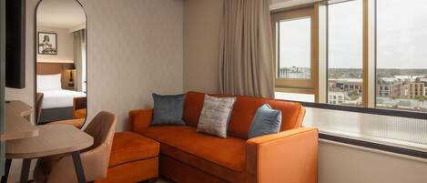 Et moderne hotelværelse med en sofa, et skrivebord og et stort vindue med udsigt over byen.