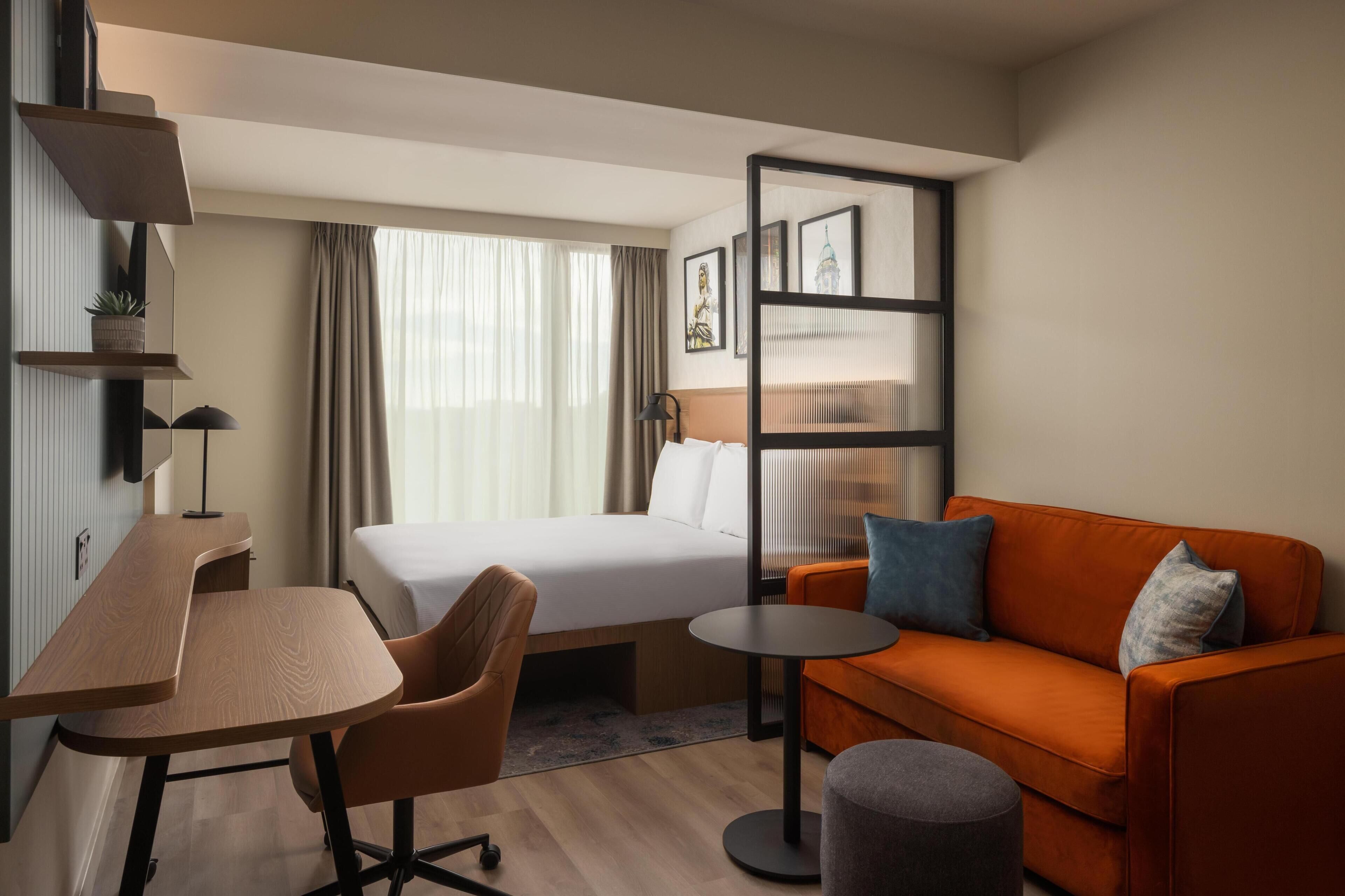 Una habitación de hotel con cama, escritorio, sofá, una mesita y un estante en la pared.