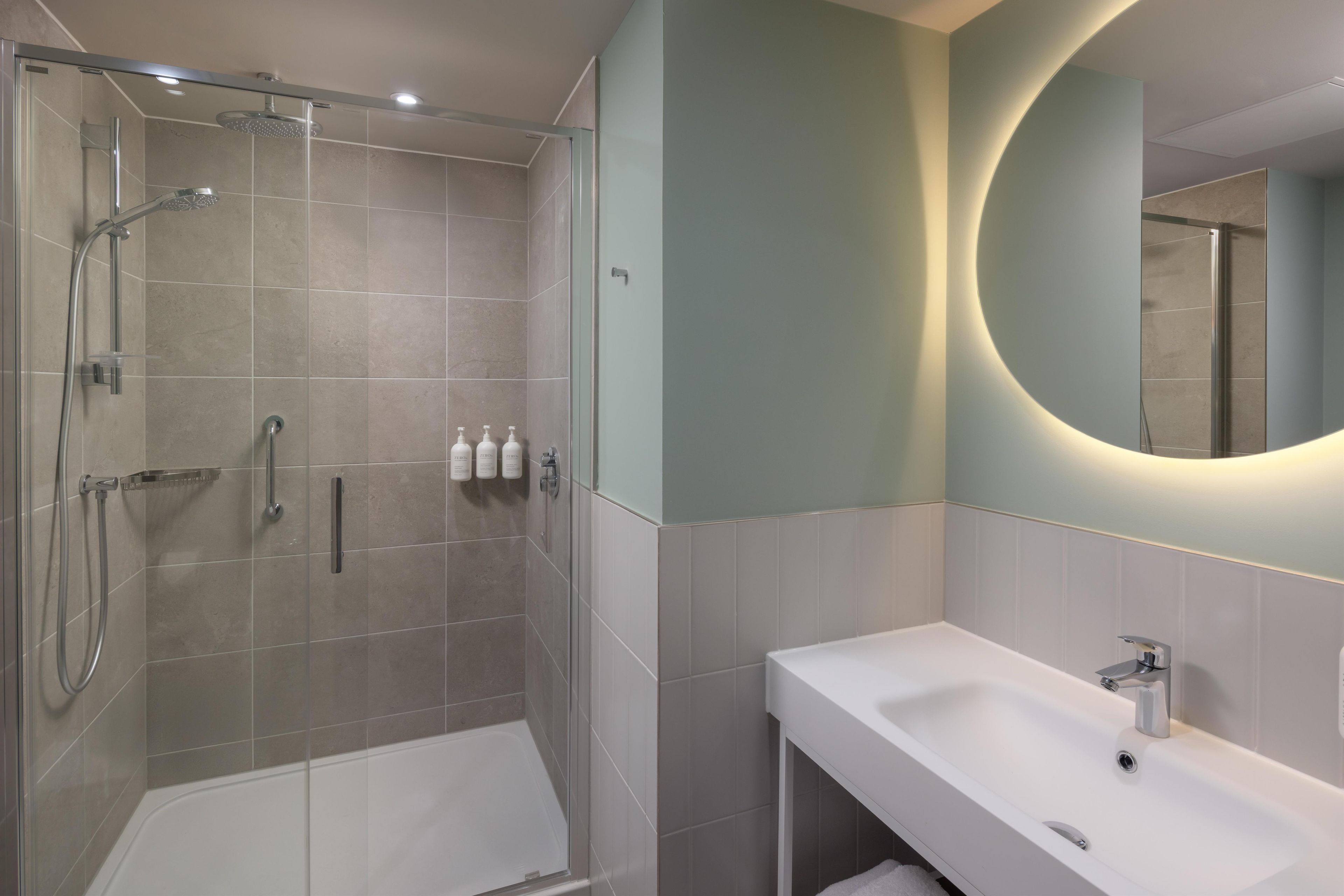 A modern bathroom with a glass-enclosed shower, a white sink, and a mirror with a circular light.