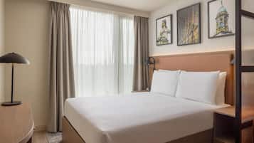 Habitación de hotel con una cama grande, una mesita de noche con lámpara, una mesita auxiliar y un televisor montado en la pared.