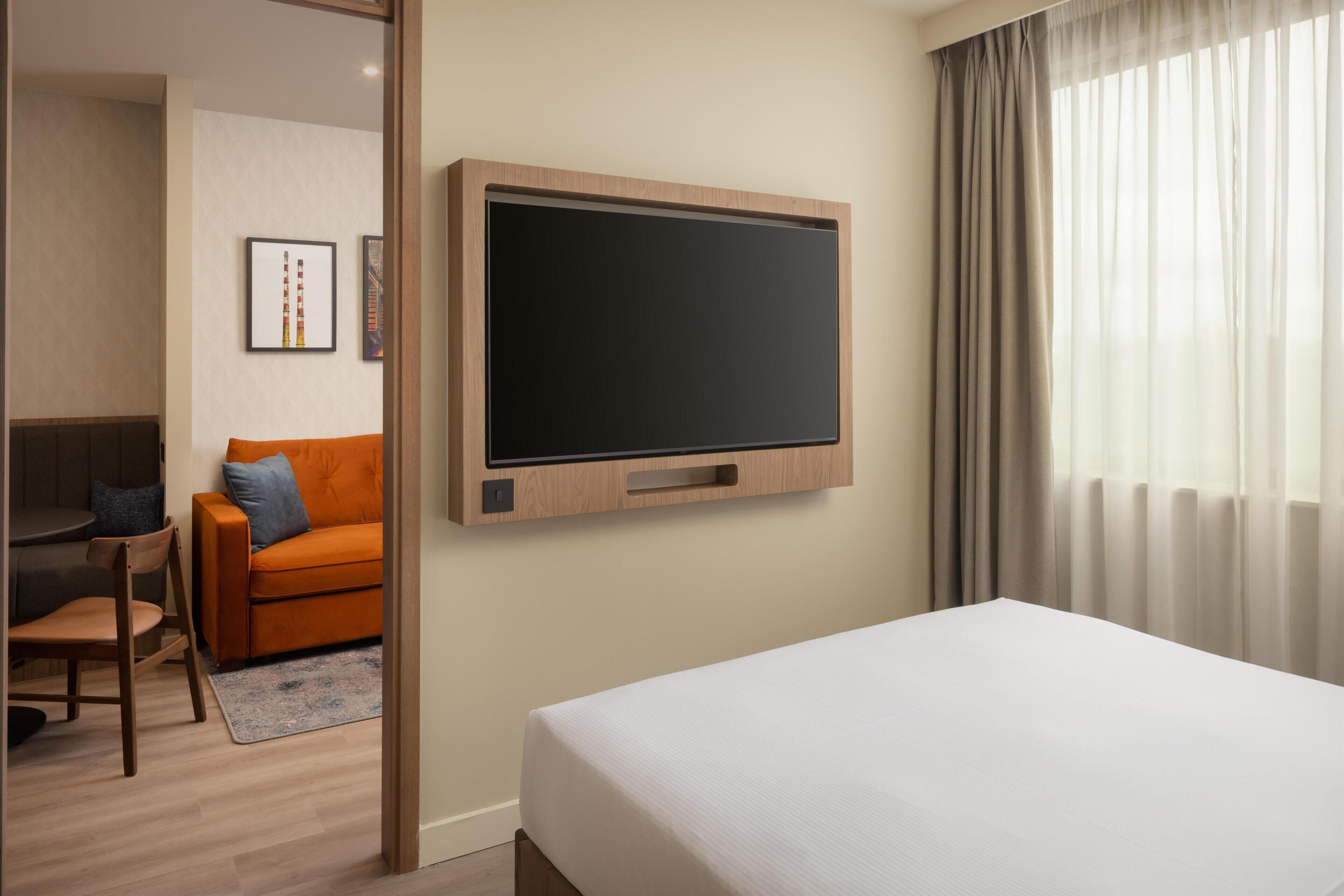 A hotel room with a large flat-screen TV mounted on the wall, a bed, a sofa, a chair, and a desk.