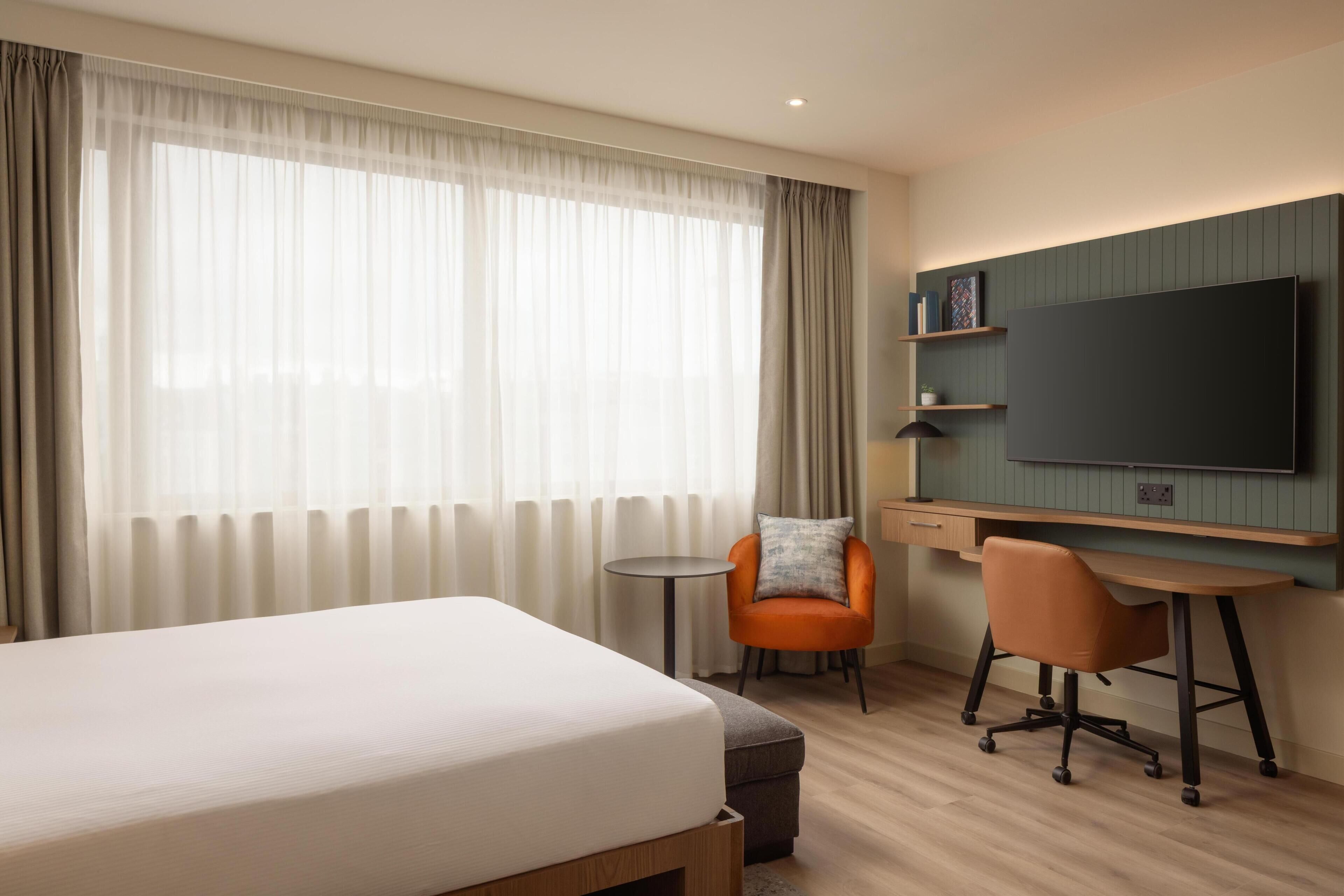 Habitación de hotel con cama, escritorio, silla, televisor y ventana con cortinas.