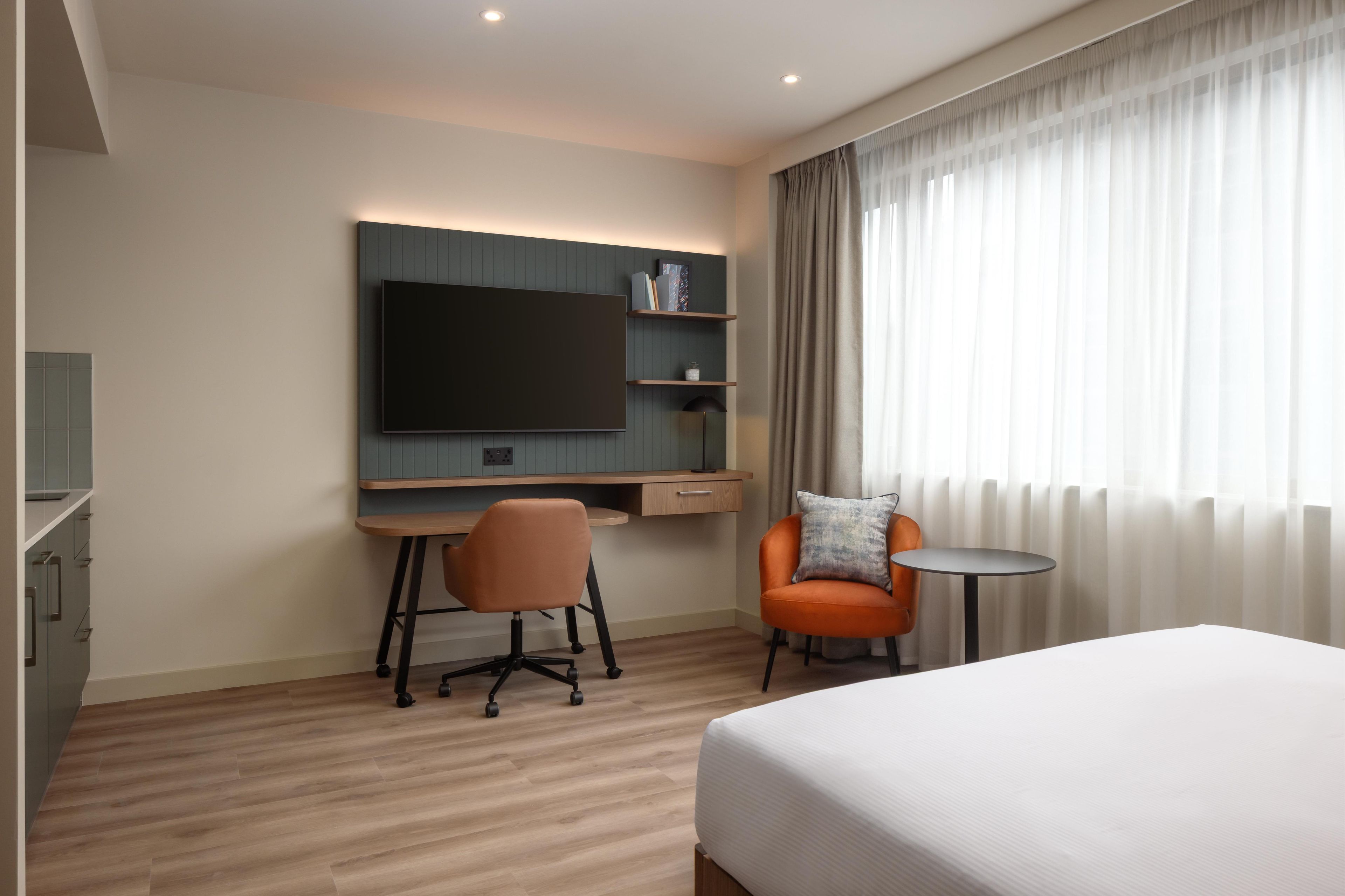 A modern hotel room with a large bed, a desk with a chair, a television, and a small table.