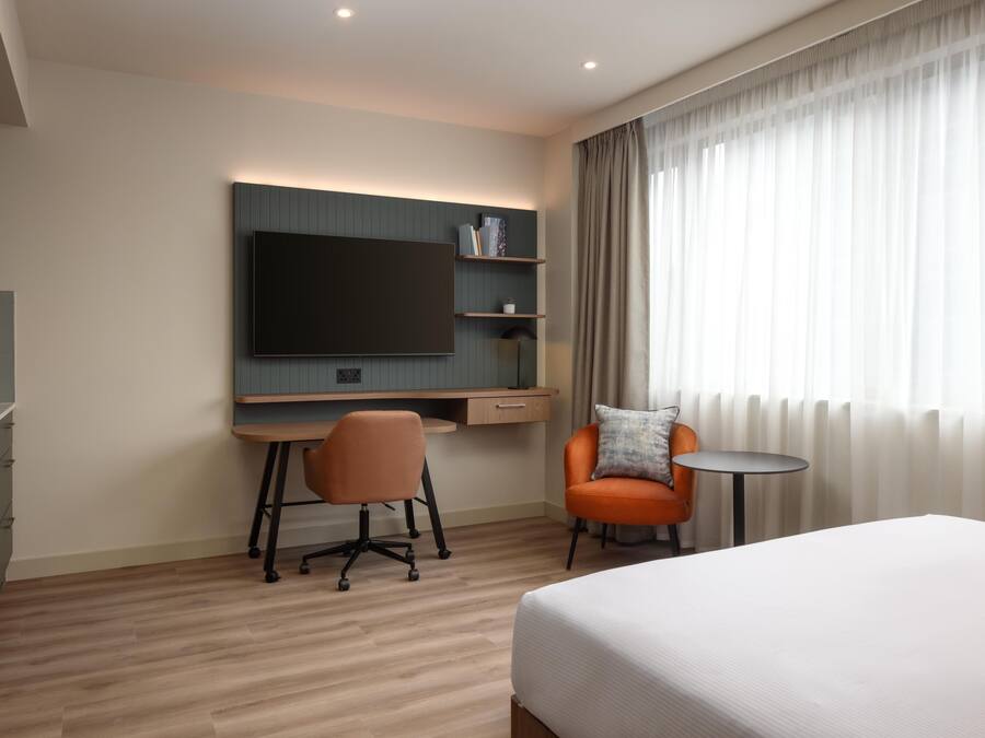 A modern hotel room with a large bed, a desk with a chair, a television, and a small table.