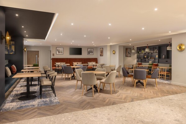 Un comedor moderno con varias mesas y sillas, suelo de madera con diseño de espiga y luces empotradas en el techo.