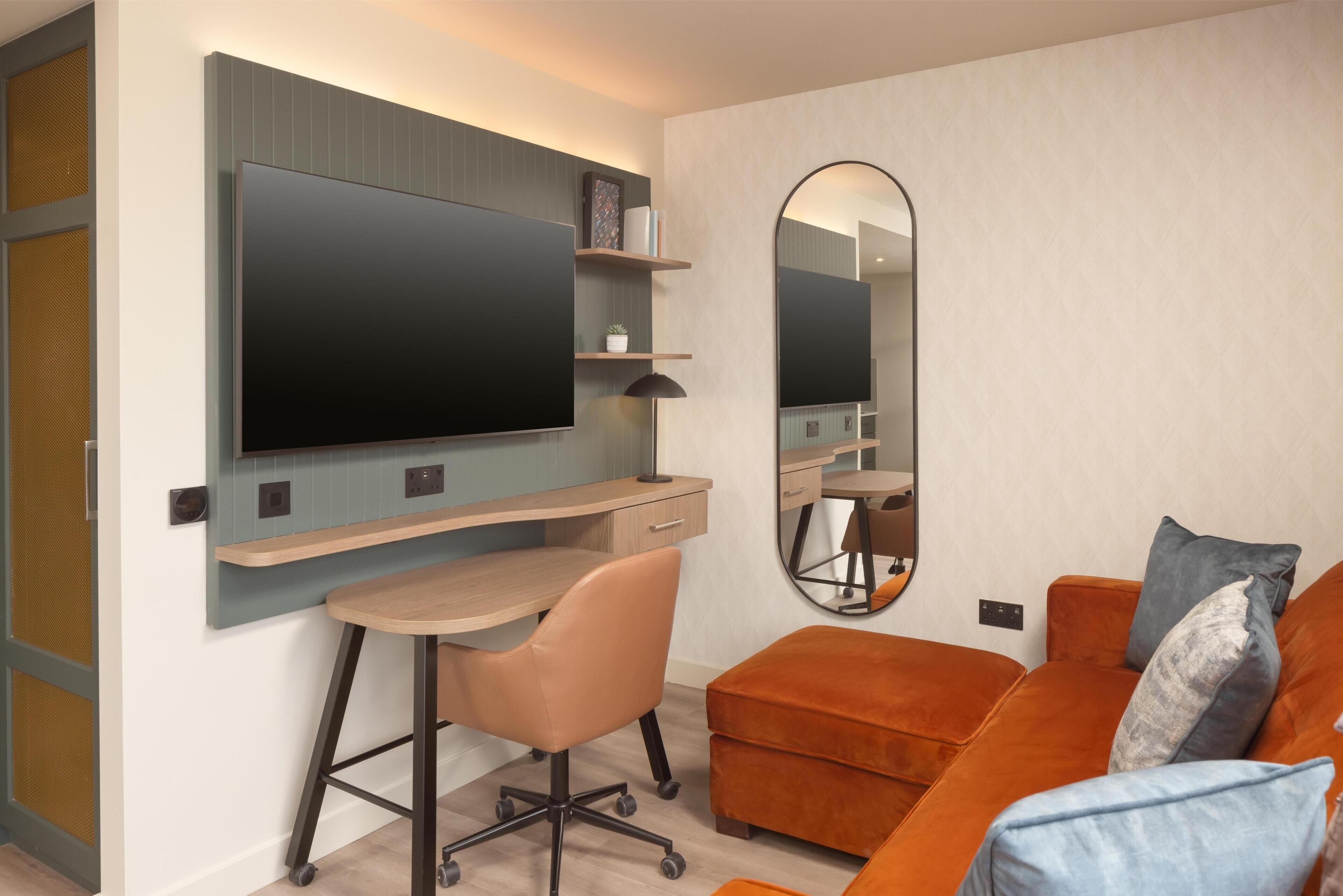 Une chambre moderne avec un téléviseur fixé au mur, un bureau avec une chaise, un canapé avec des coussins et un grand miroir.