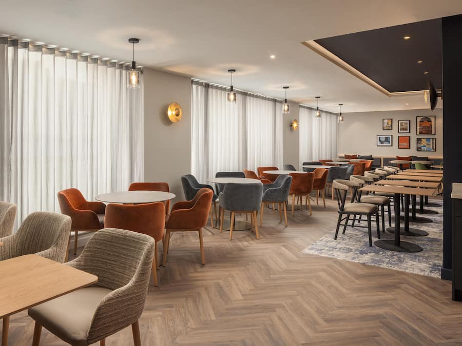 A modern hotel lobby with wooden flooring, round tables, and a mix of upholstered chairs.