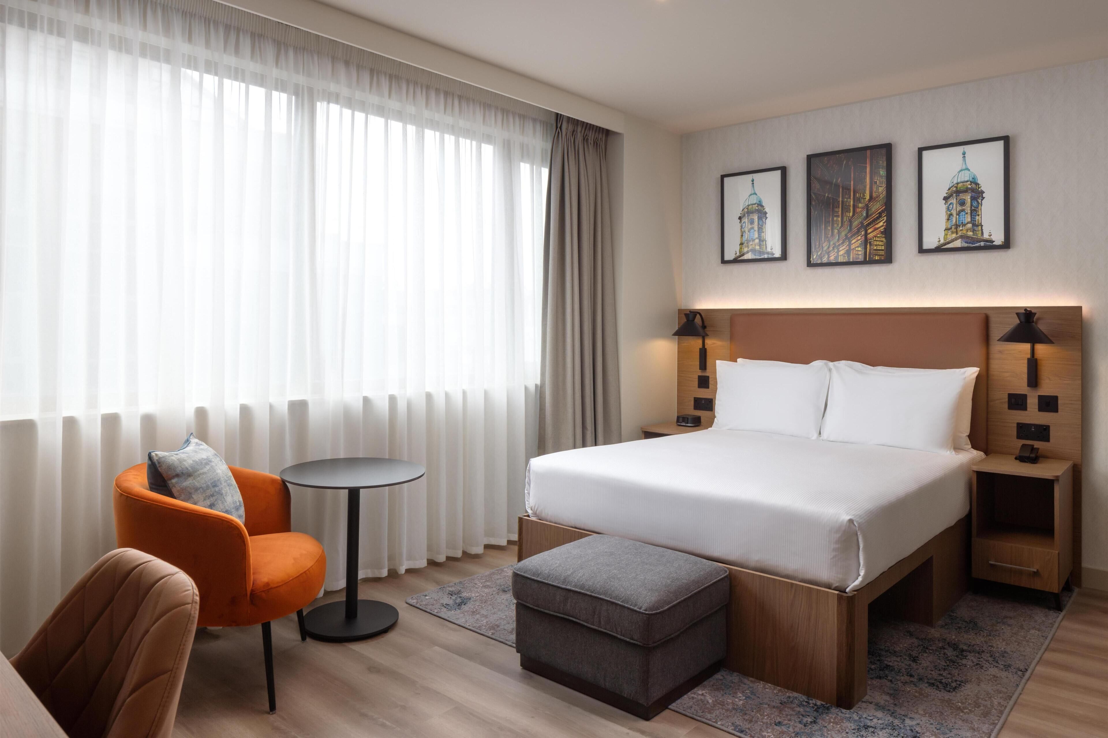 Une chambre d’hôtel avec un grand lit, une petite table ronde, une chaise orange, une table de chevet en bois et un pouf gris.