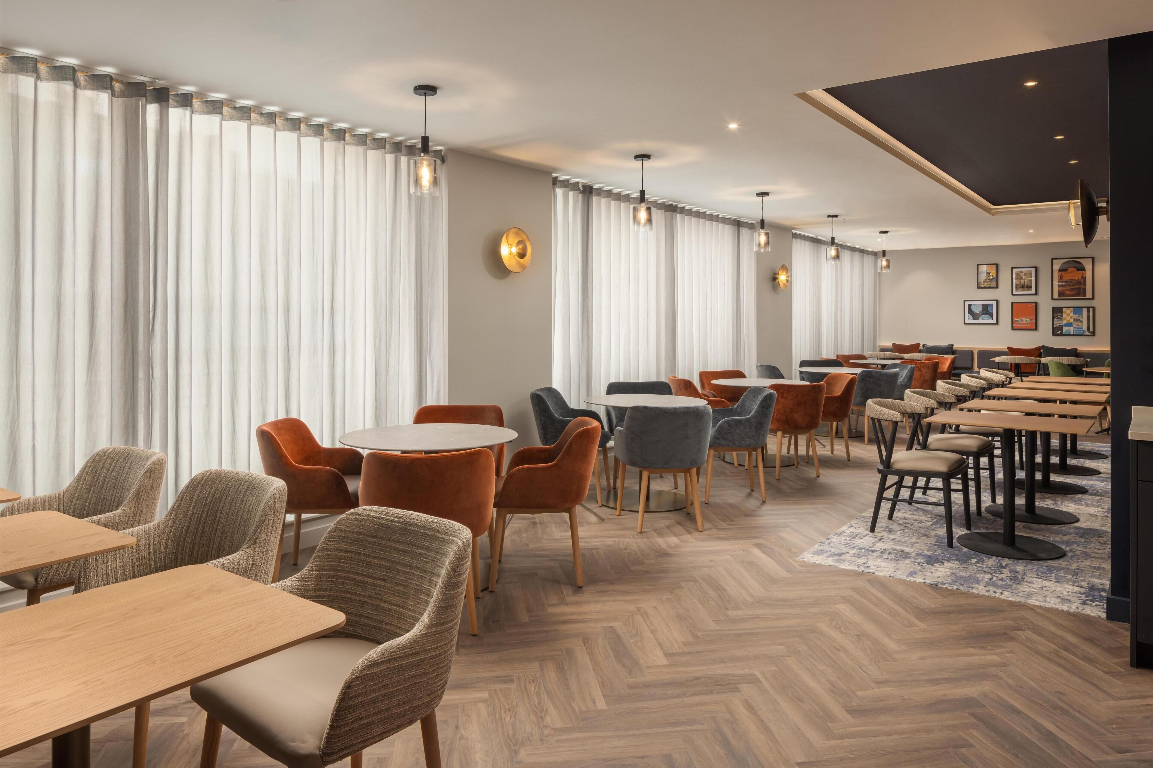A modern hotel lobby with wooden flooring, round tables, and a mix of upholstered chairs.