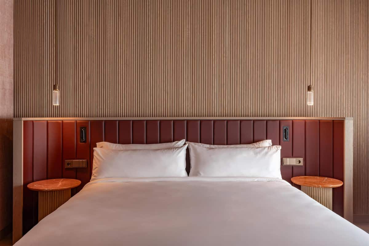 Una habitación de hotel con una cama grande, dos mesitas de noche, un cabecero de madera y un panel vertical en la pared.