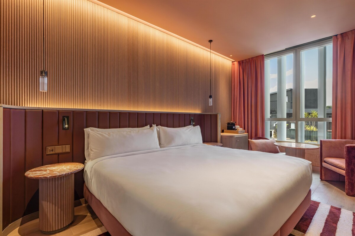 A hotel room with a large bed, a bedside table, a chair, a small table, and a large window with curtains.