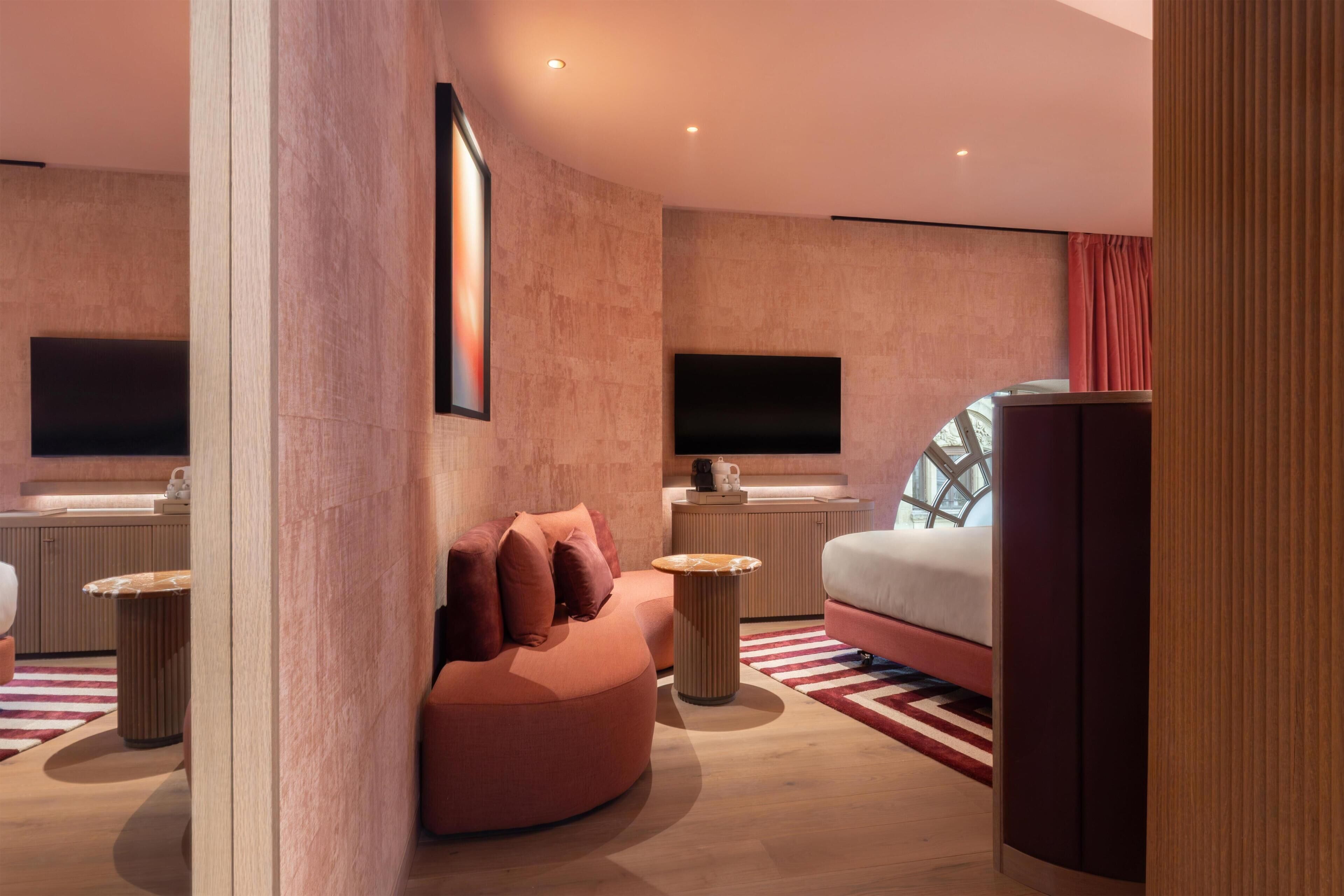 A modern hotel room with a large bed, a seating area, a TV, and a large window.