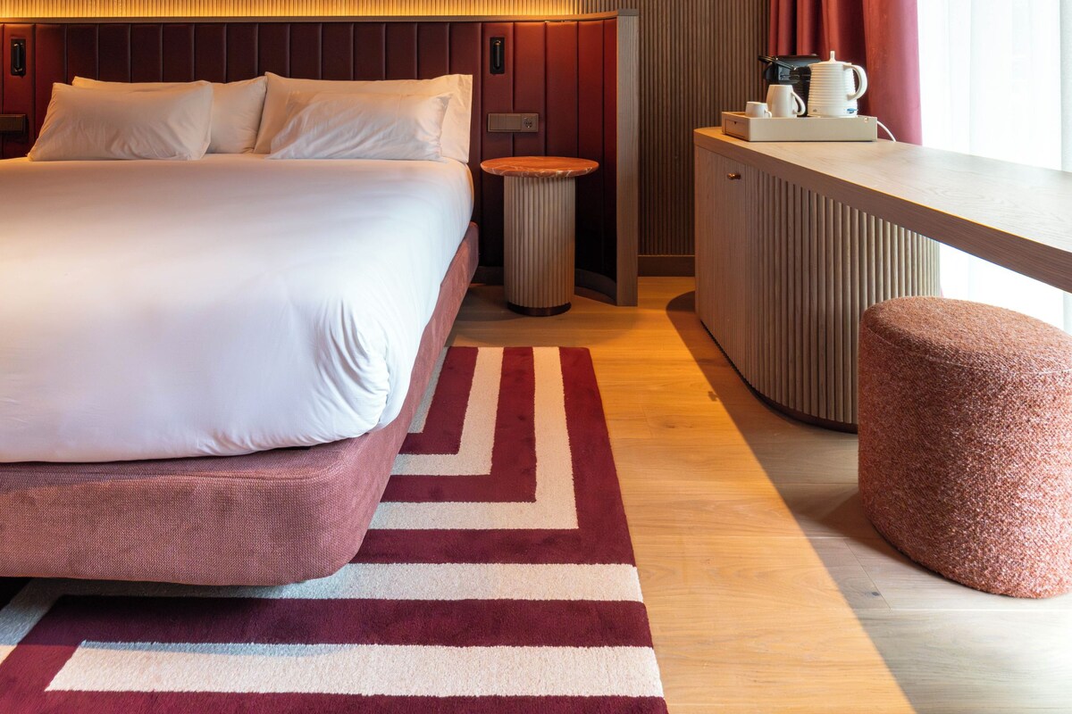 A modern hotel room with a large bed, a bedside table, a small round table, a radiator, and a window with curtains.