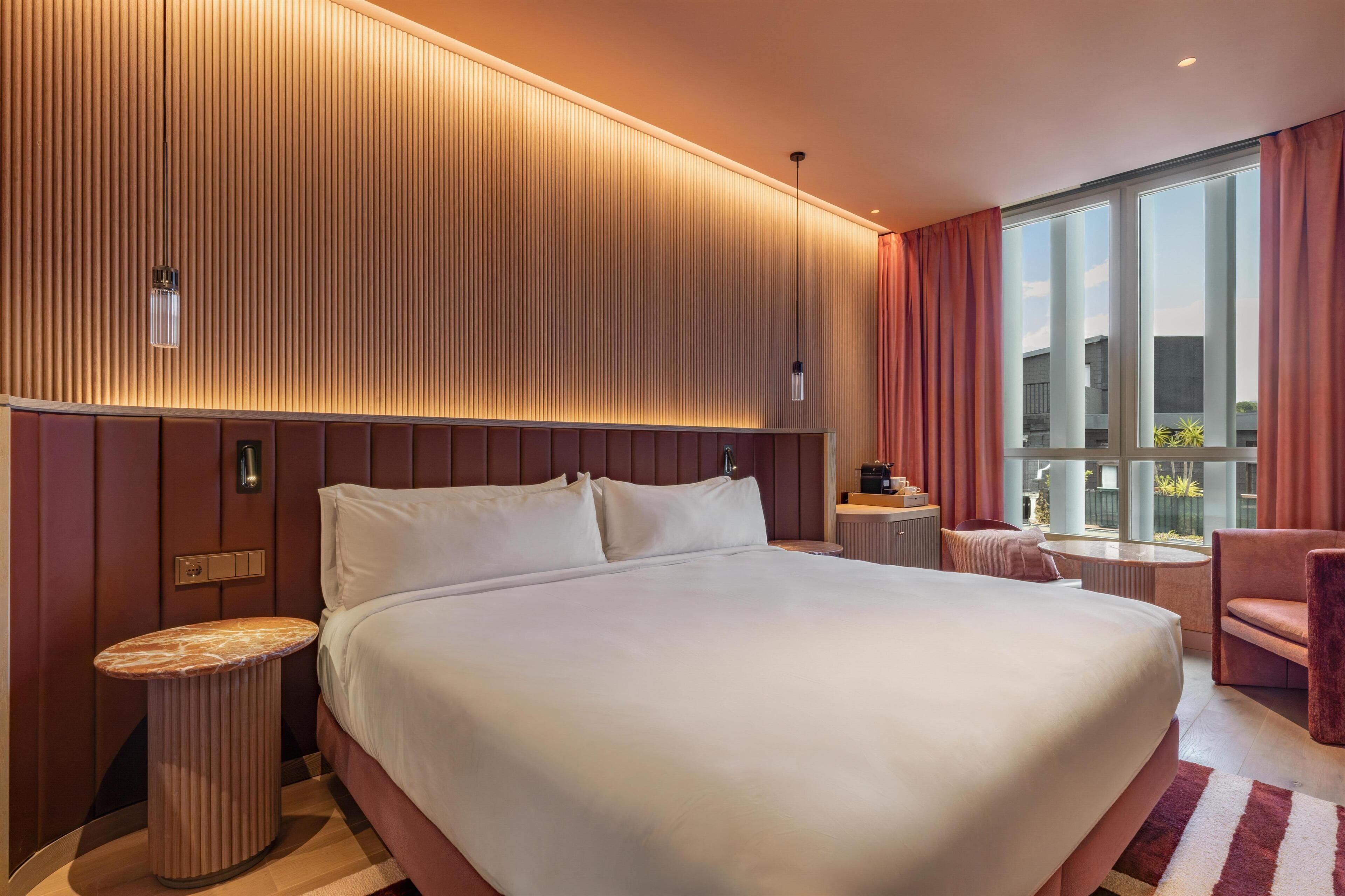 Hotelkamer met een groot bed, nachtkastje, stoel, klein tafeltje en een groot raam met gordijnen.