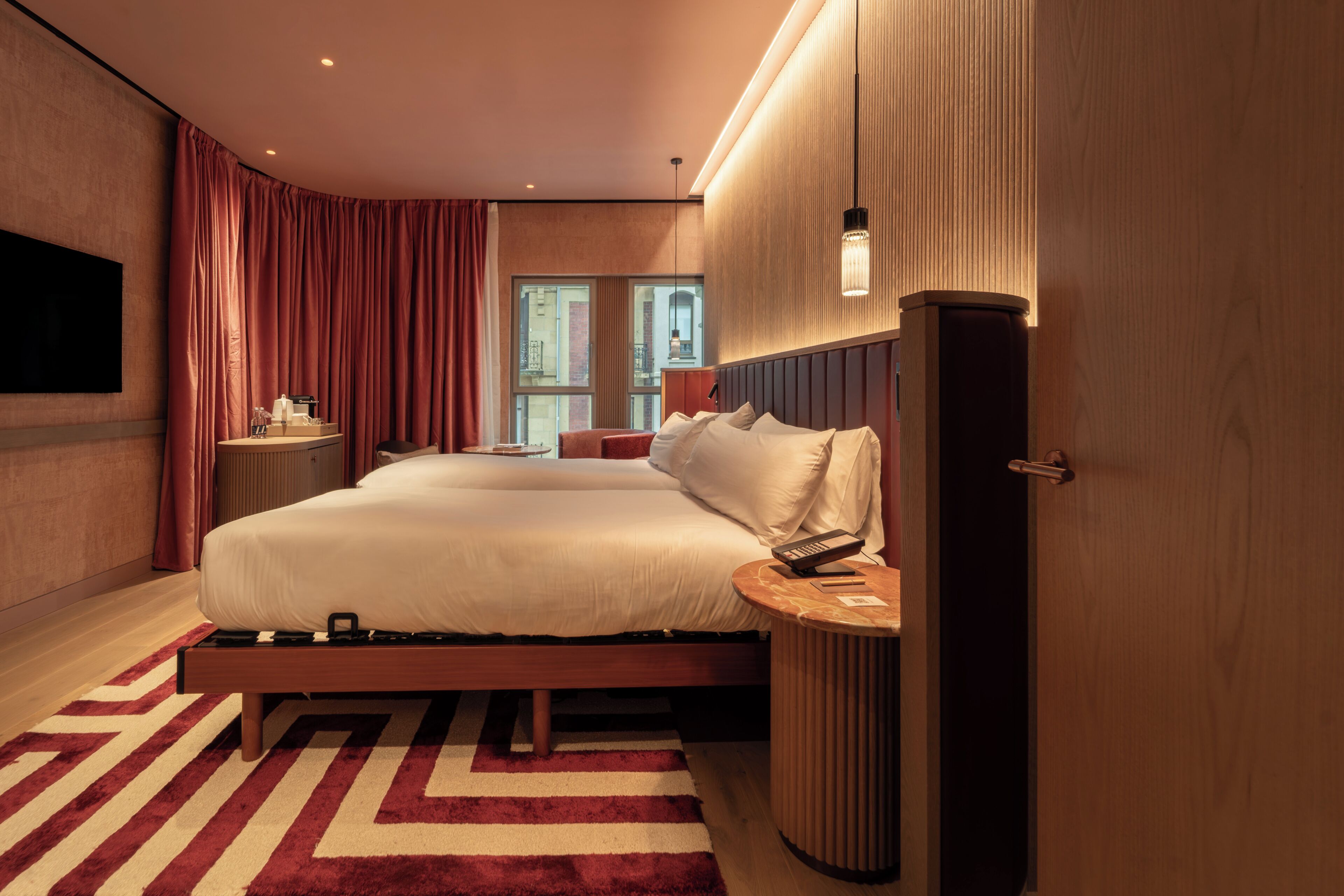 A modern hotel room with a large bed, a flat-screen TV, a bedside table, and a view of the city through the window.