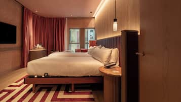 A modern hotel room with a large bed, a flat-screen TV, a bedside table, and a view of the city through the window.