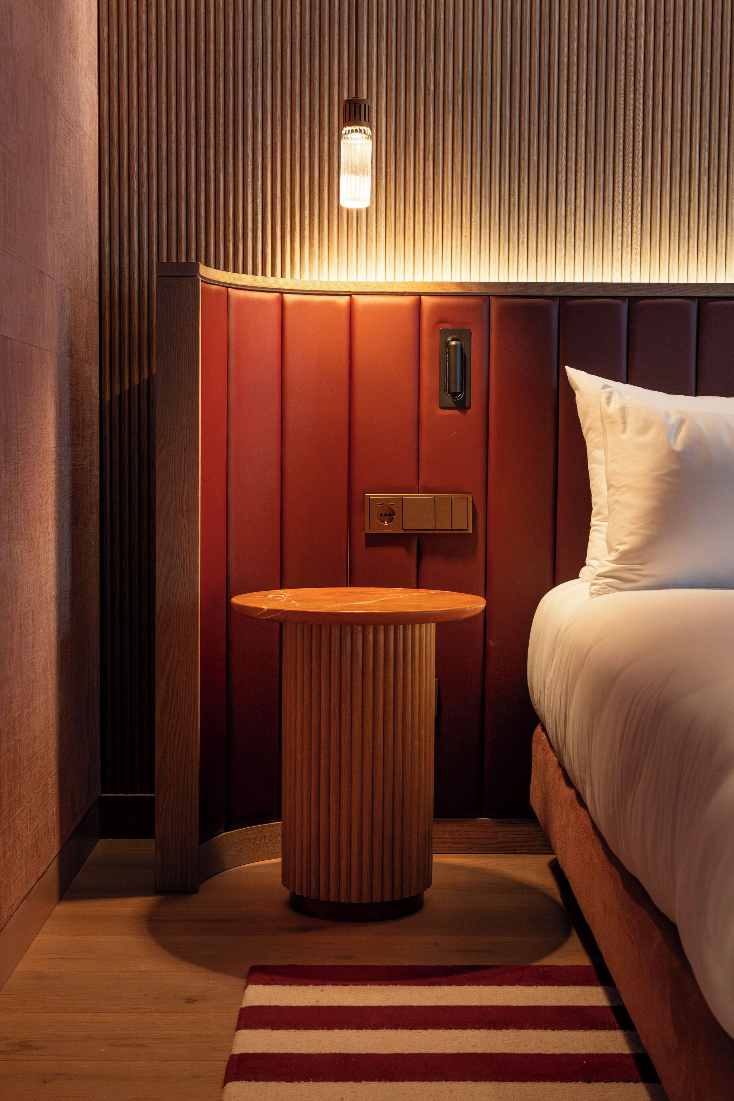A hotel room with a bed, a round wooden nightstand, a wall-mounted light, and a red-striped rug.