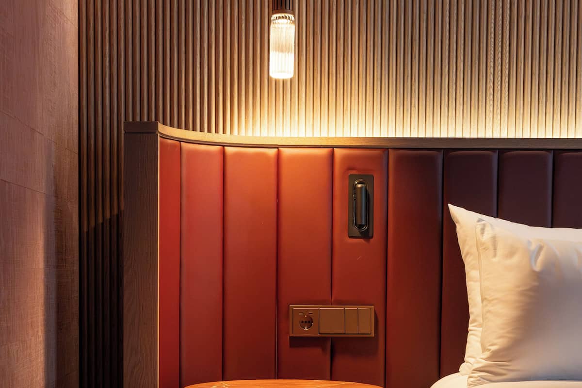 A hotel room with a bed, a round wooden nightstand, a wall-mounted light, and a red-striped rug.