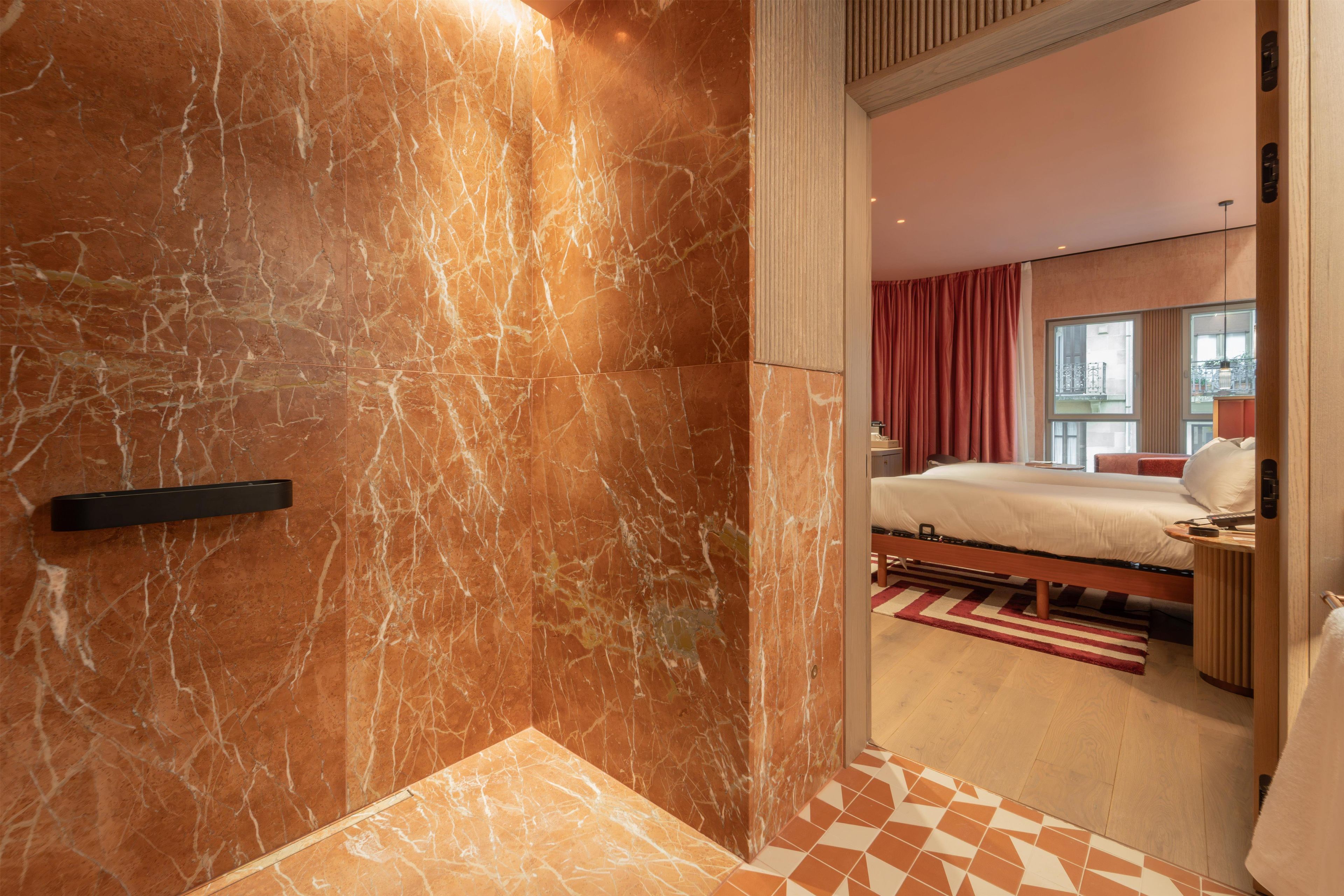 Habitación de hotel con pared de mármol, una cama, una mesita de noche y vistas de la ciudad a través del ventanal.