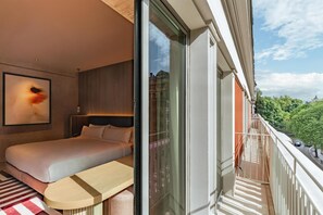 Een moderne hotelkamer met een bed, een klein tafeltje, een groot raam en een balkon met uitzicht op groen en een gebouw in de verte.