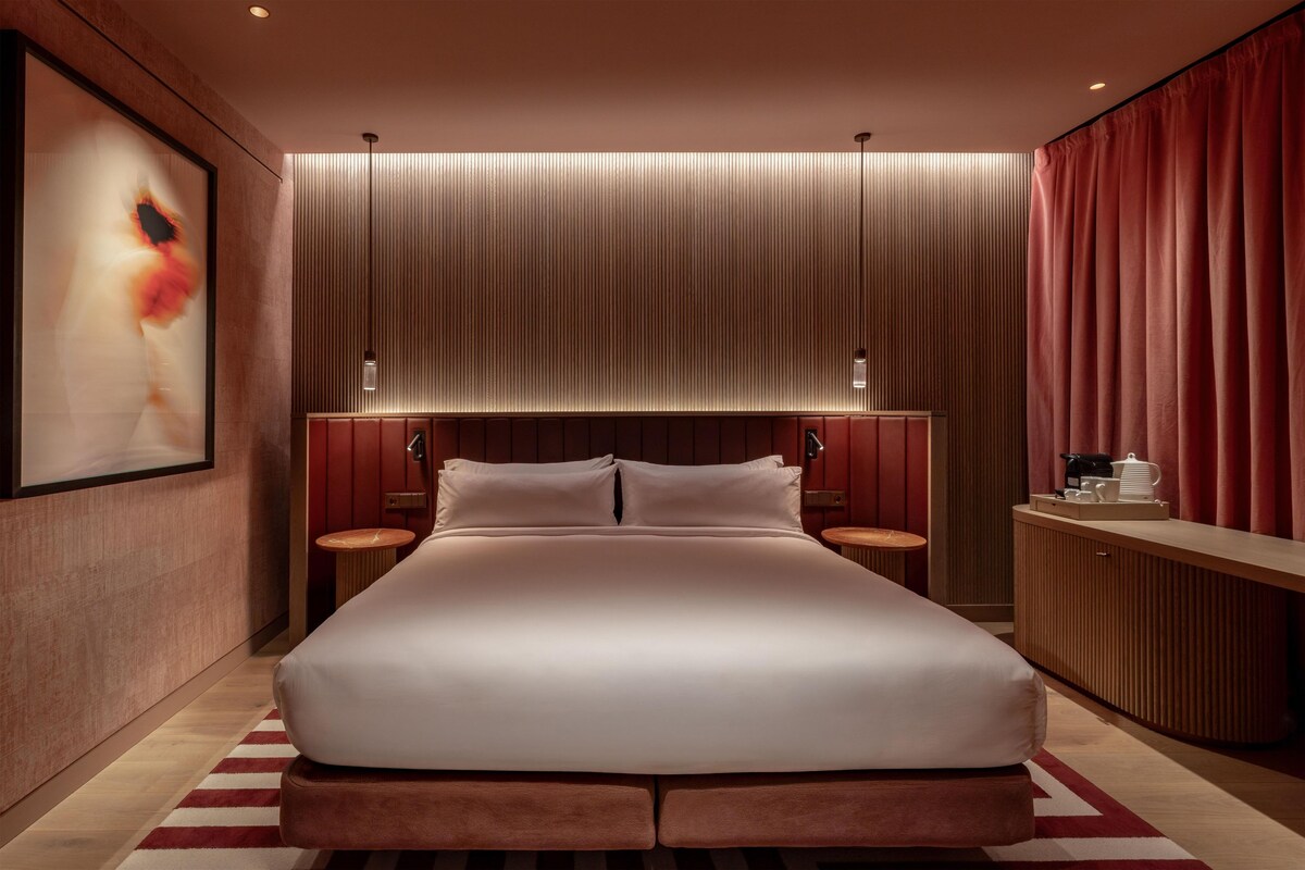 A modern hotel room with a large bed, wooden headboard, two bedside tables, and a red curtain.