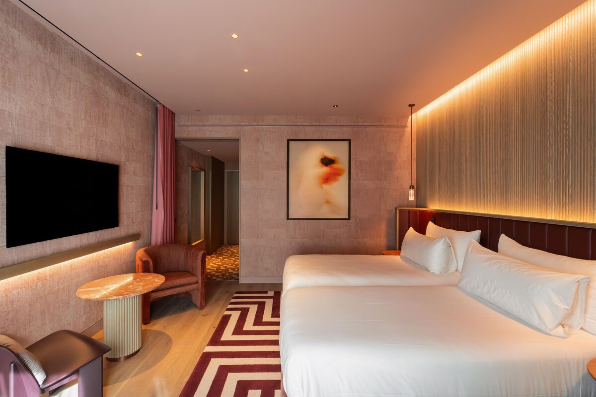 A hotel room with a large bed, a flat-screen TV mounted on the wall, a seating area with a chair and a small table, and a patterned rug on the floor.
