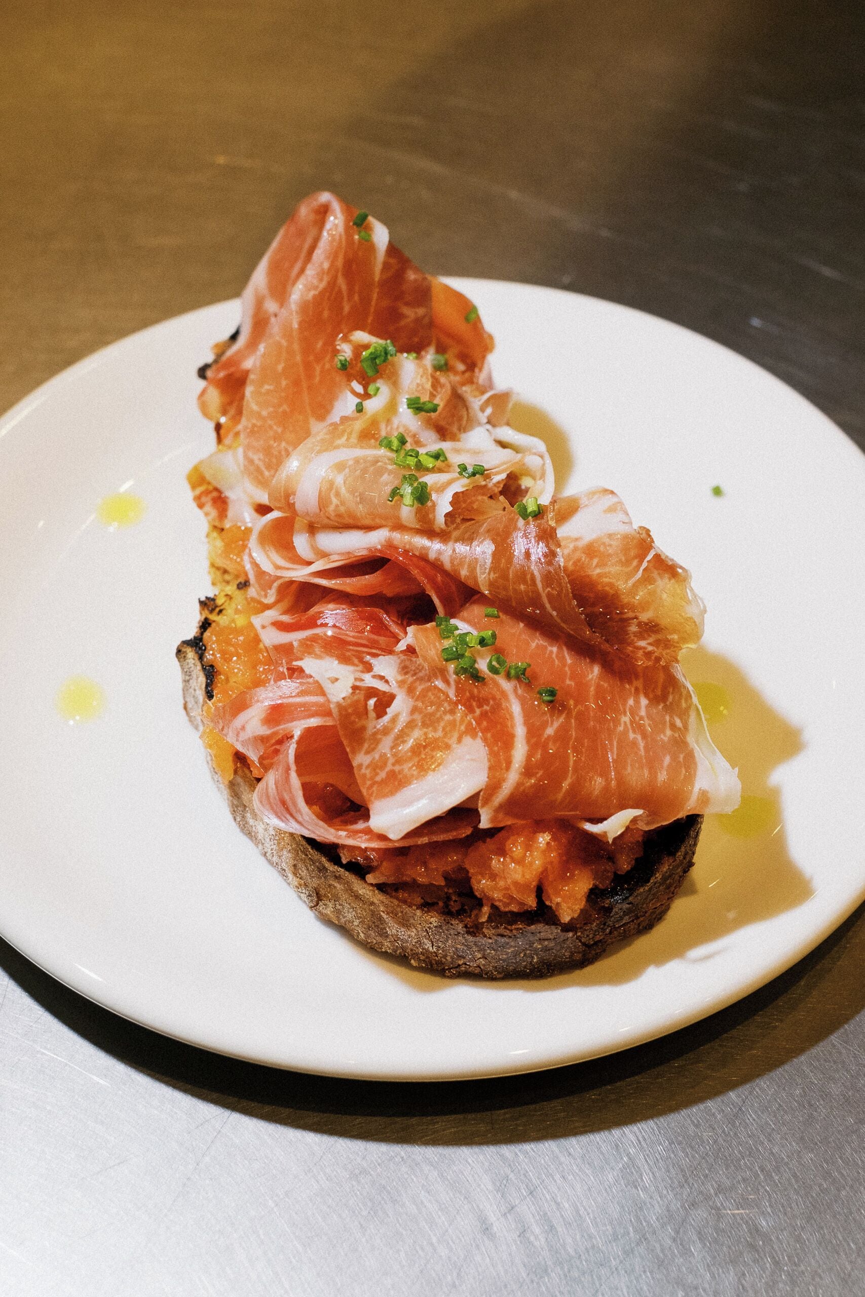 Un plato con una rebanada de pan cubierta con una loncha de embutido y un chorrito de aceite de oliva.