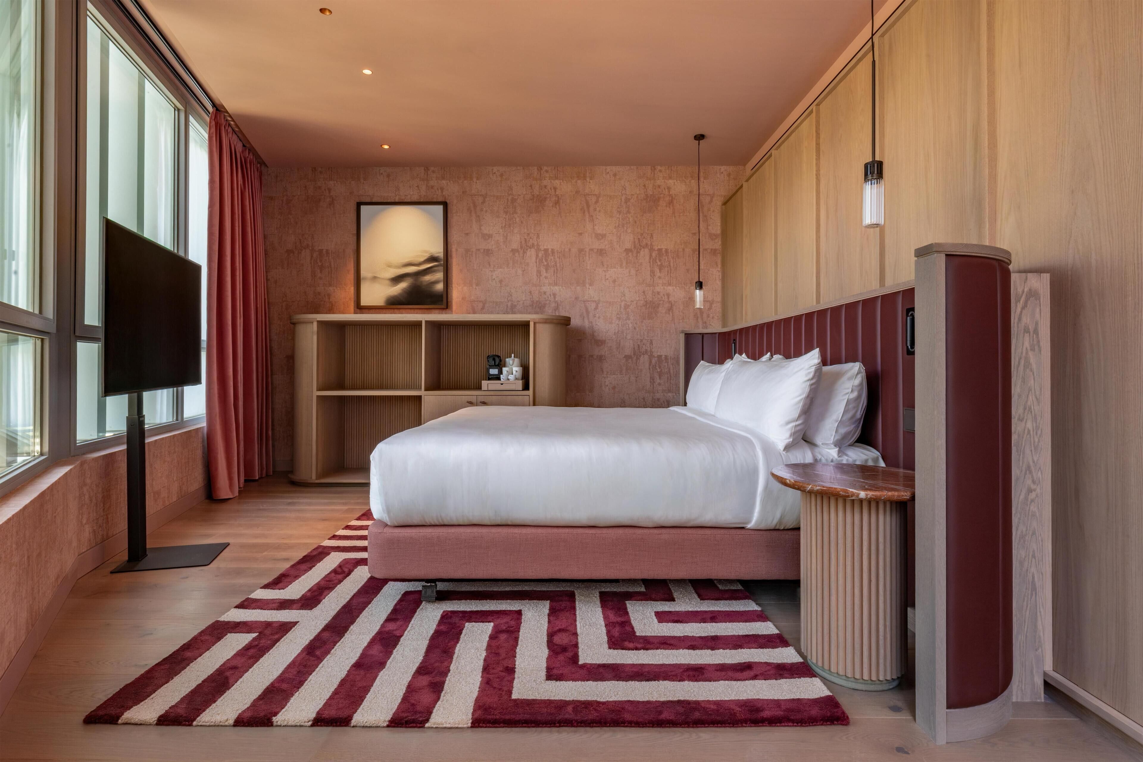 Une chambre d’hôtel moderne avec un grand lit, une télévision, une tête de lit en bois, un tapis rouge et blanc, et une bibliothèque.