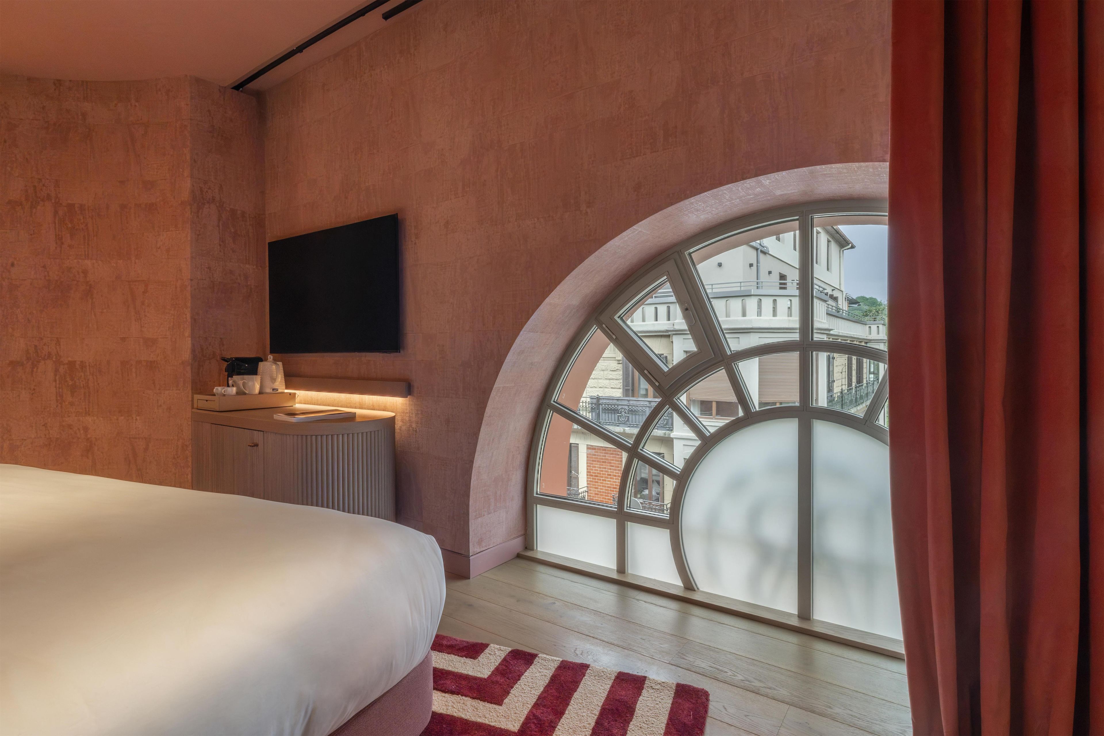 Un dormitorio con un gran ventanal arqueado, una cama, una mesita de noche y un televisor de pantalla plana montado en la pared.